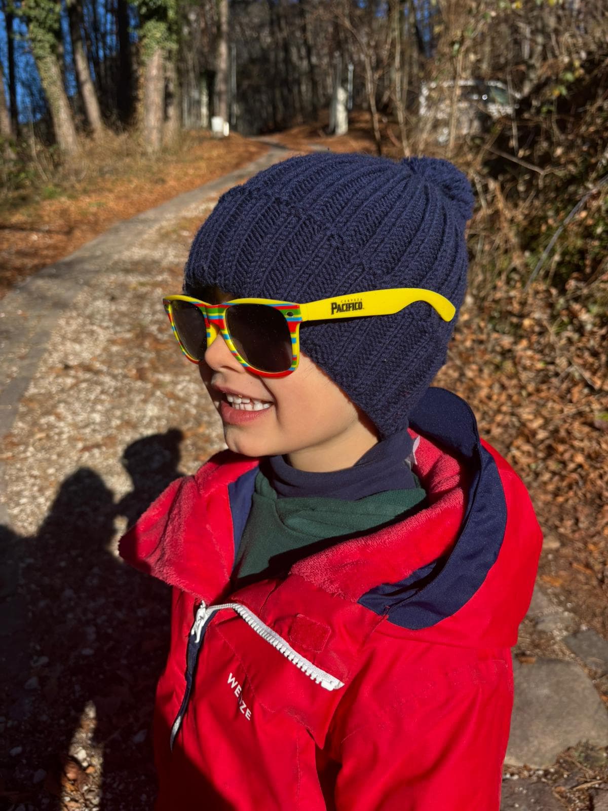 A young boy in a navy beanie and Pacífico-branded sunglasses, smiling, on a leaf-strewn path in late autumn light