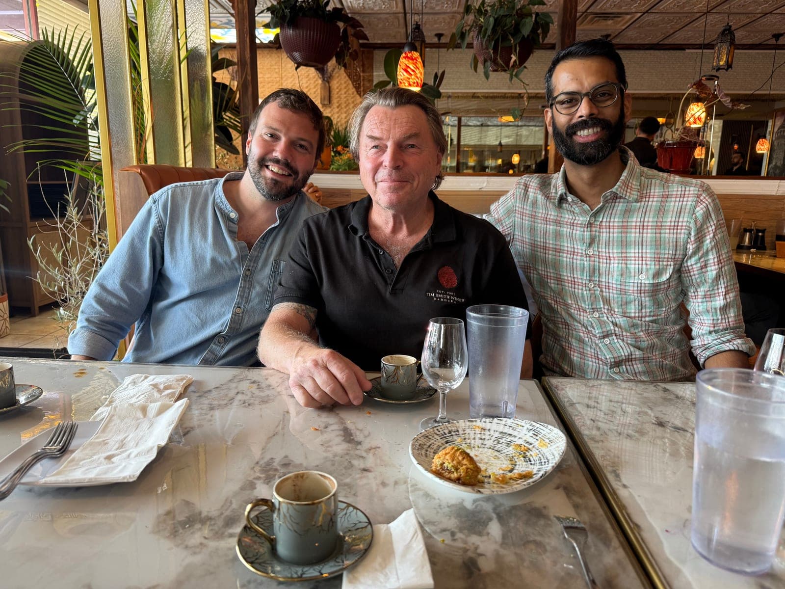 Three men smiling at the camera across a marble restaurant table after lunch, Tim Smith in the middle in a black Tim Smith Wines polo, with Turkish coffee cups, a water glass and a plate of baklava crumbs in front of them.